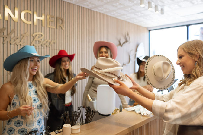 Friends smiling while shopping for colorful cowboy hats at a bright western hat boutique, exchanging a beige hat with a braided band and feather detail at the counter.