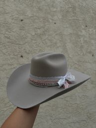 Light gray felt wide-brim cowboy hat with pink ribbon, lace and beaded trim tied in a bow, held against a textured stucco wall