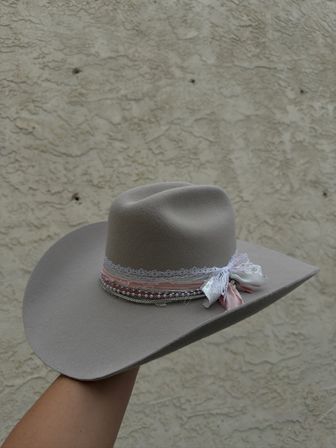Light gray felt wide-brim cowboy hat with pink ribbon, lace and beaded trim tied in a bow, held against a textured stucco wall