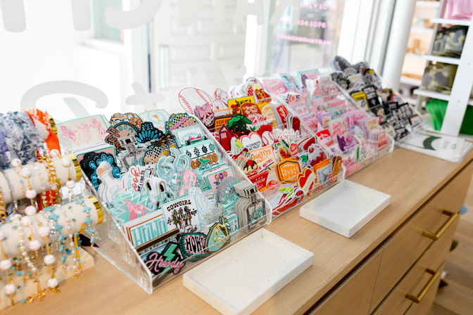 Playful boutique accessory display of colorful decorative patches and stickers in clear acrylic racks on a wooden counter, with beaded bracelets on a nearby stand