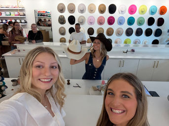 Three smiling women taking a selfie inside a bright hat boutique with a colorful wall display of cowboy hats; one woman behind the counter holds a white cowboy hat while wearing a dark brown one.