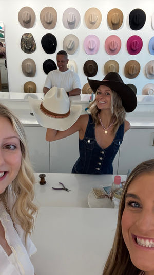 Three smiling women taking a selfie inside a bright hat boutique with a colorful wall display of cowboy hats; one woman behind the counter holds a white cowboy hat while wearing a dark brown one.