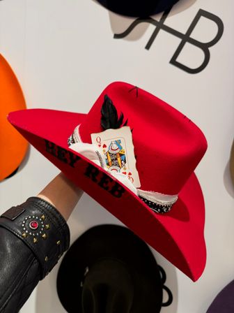 Hand holding bright red felt cowboy hat with Queen of Hearts playing card tucked into a bandana-trim hatband and a black feather, displayed on a hat shop wall.