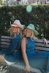 Two smiling women in denim outfits and pastel cowboy hats sitting back-to-back on a wooden bench in front of a leafy hedge on a sunny day