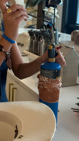 Close-up of hands carefully using a blue handheld torch to shape a white felt hat brim in a hat-making workshop, with metal tools, jars of brushes and stacked hats on the counter.