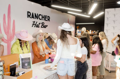 Group of women shopping and trying on cowboy hats at a pink western-style hat bar boutique, lively counter scene with neon lights and cactus wall art.