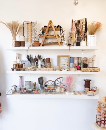 Bright boho craft studio wall shelves neatly arranged with ribbons, spools, beads, trims, feathers and dried grasses for DIY projects.