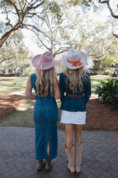 Two friends standing with their backs to the camera in a sunlit park, wearing denim outfits, pastel cowboy hats with ribbons and western boots beneath sprawling oak trees