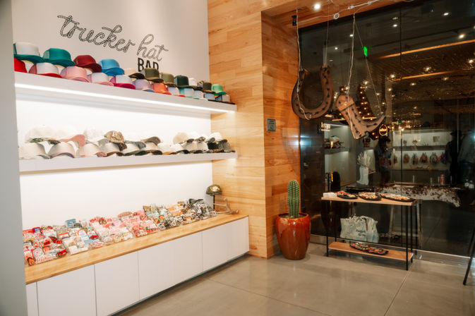Colorful trucker hat display in a modern boutique — illuminated white shelves stacked with multicolored caps and embroidered patches on a wooden counter, tall potted cactus and glass storefront with hanging leather horseshoes.