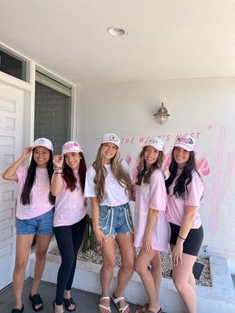 Five smiling women in matching pink t-shirts and white-pink trucker hats posing on a sunny front porch in front of a pastel pink cactus mural