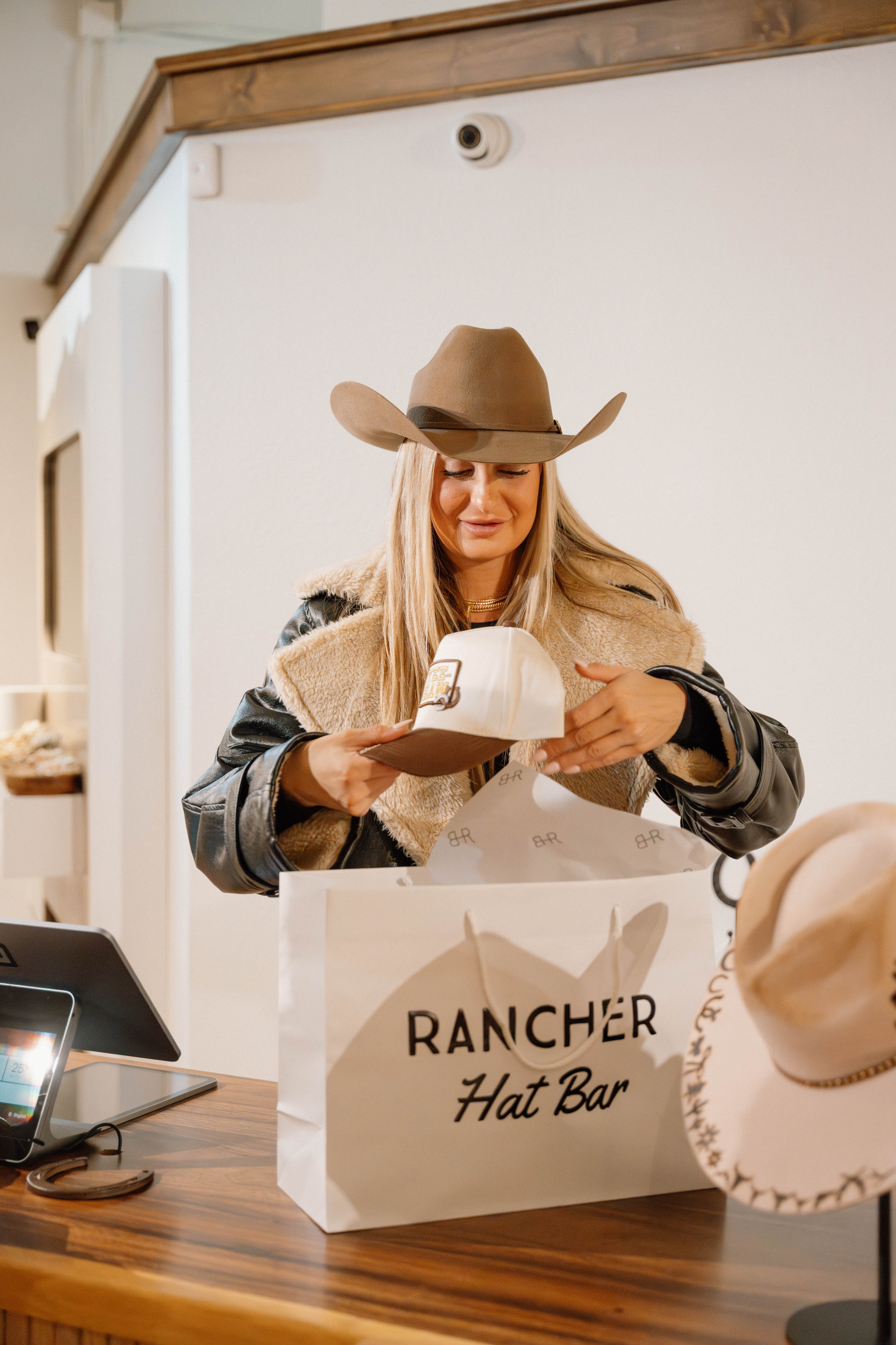 Smiling shopper wearing a cowboy hat and shearling jacket at a western hat boutique checkout, lifting a brown-and-white trucker cap from a white shopping bag with a hat-shop logo, felt hats and register on the wooden counter.