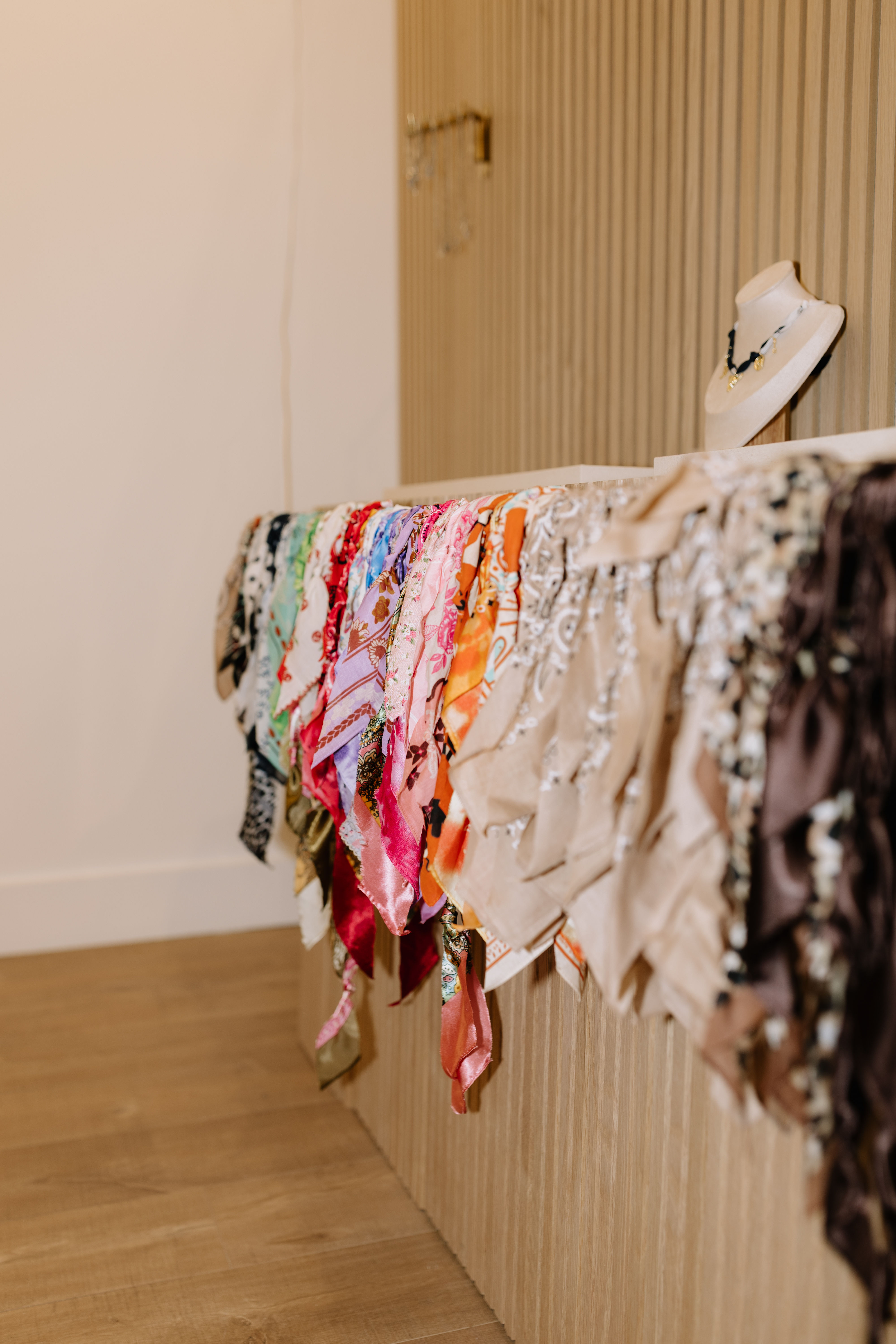 Vibrant patterned silk scarves cascading over a corrugated wooden counter in a boutique display, with a necklace-styled bust on top and warm hardwood floor beneath.
