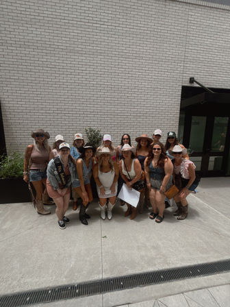 Group of women in cowgirl outfits—cowboy hats, boots and summer dresses—posing for a bachelorette party photo in a sunny urban courtyard against a white brick wall.