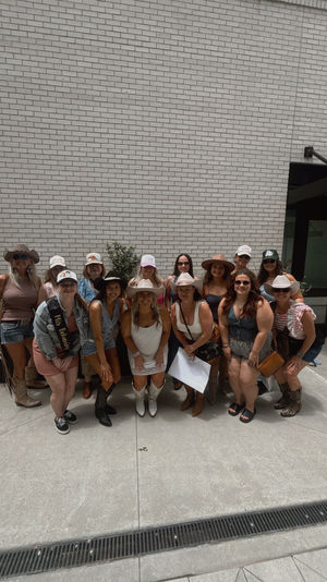 Group of women in cowgirl outfits—cowboy hats, boots and summer dresses—posing for a bachelorette party photo in a sunny urban courtyard against a white brick wall.