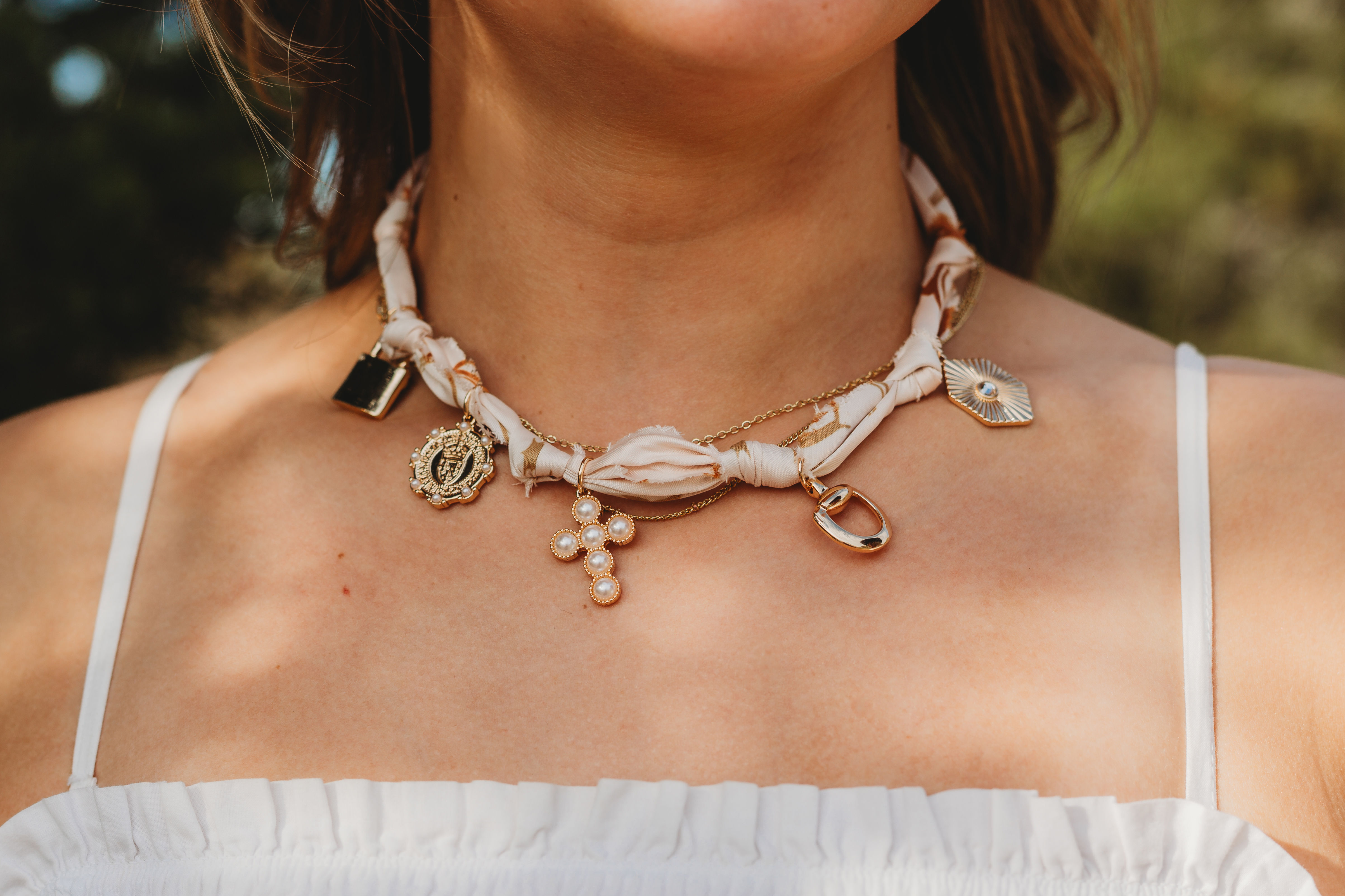 Close-up of a woman's neck wearing a twisted silk choker layered with gold charms — a pearl cross pendant, medallion and geometric charms — styled with a white spaghetti-strap summer top, outdoor fashion accessory.