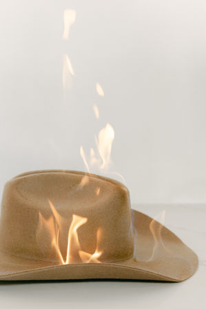 Close-up of a tan felt cowboy hat burning, with flickering orange flames and wisps of smoke rising against a plain white background.