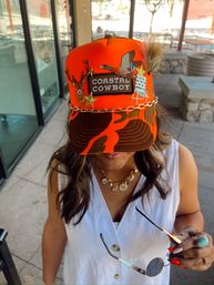 Bright orange trucker hat reading Coastal Cowboy with embroidered deer, flying duck, gold stars, chain trim and faux fur tuft, worn by a person in a white button dress holding round sunglasses on an outdoor patio.
