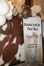 Western-themed display with a vertical sign for a hat bar and custom/trucker hats, model in a cowboy hat, cowhide wall, decorative horseshoe and beige-brown balloon garland