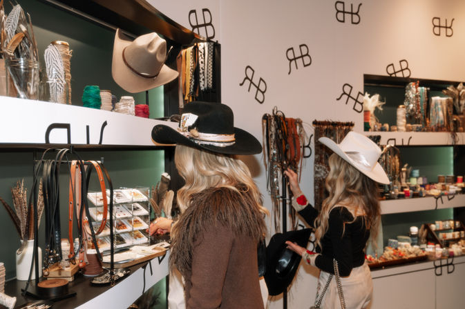 Two women in cowboy hats browse a western accessories boutique, inspecting leather belts, hat displays, feathers, jewelry and small gift items on lit wall shelves.