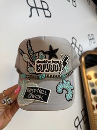 Gray western trucker hat with patches reading "should've been a COWBOY" and "RAISE HELL, COWGIRL", turquoise floral embroidery, beaded rope trim and star studs, held by a hand with pink nails.