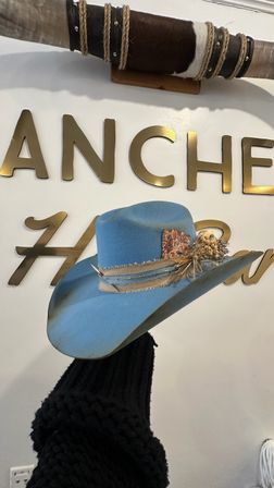 Sky-blue felt cowboy hat with frayed leather band, feather and dried flowers, displayed on a mannequin against a white wall with gold letters and a mounted longhorn in a western shop display.