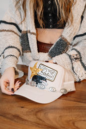 Close-up of a pink boho trucker hat on a wooden table — gold glitter star, "Ain't my first Rodeo" patch and silver concho trim — hands in a chunky knit cardigan adjusting the hat