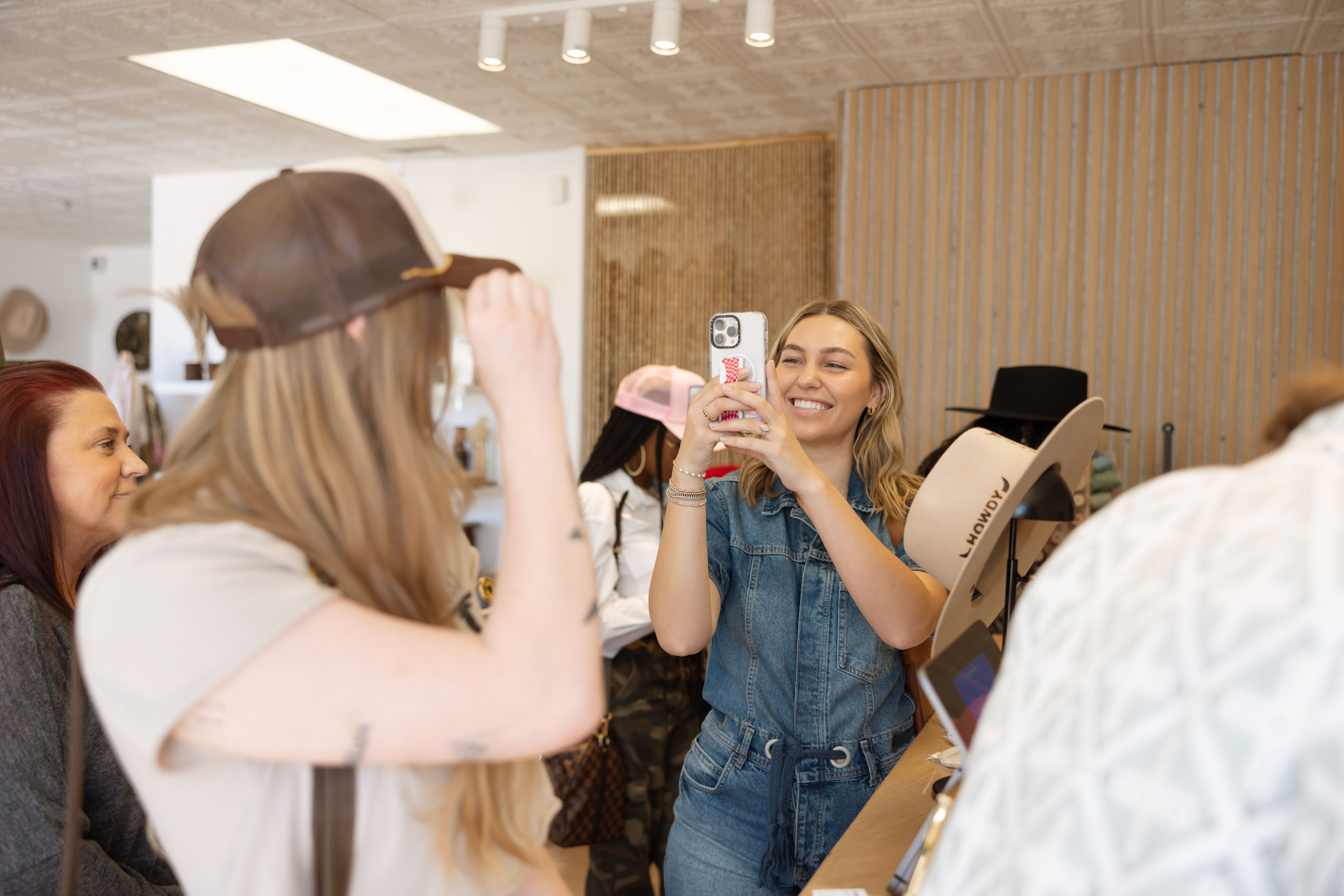Happy woman in denim taking a phone photo of a friend trying on a baseball cap inside a bright hat boutique with other shoppers
