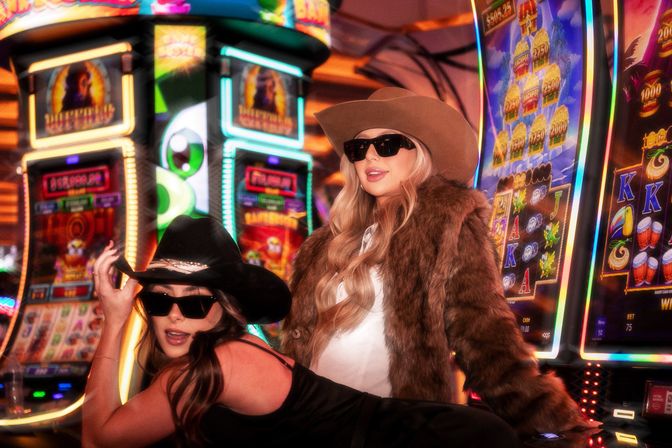 Two people posing on a neon-lit casino gaming floor by colorful slot machines, dressed in glamorous cowgirl style with cowboy hats, sunglasses and a faux-fur coat.