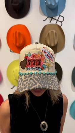Beige baseball cap with leopard-print brim, turquoise beads and embroidered Las Vegas '777' slot machine patch, worn in front of a colorful cowboy-hat wall display