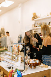 Bright boutique display of western-style concho belts, necklaces, colorful scarves and bracelets on a counter while two shoppers browse, one trying on a white cowboy hat.