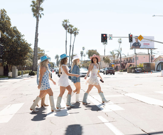 Four women in summer dresses and cowboy boots crossing a sunny coastal beach town crosswalk with palm trees and traffic lights.