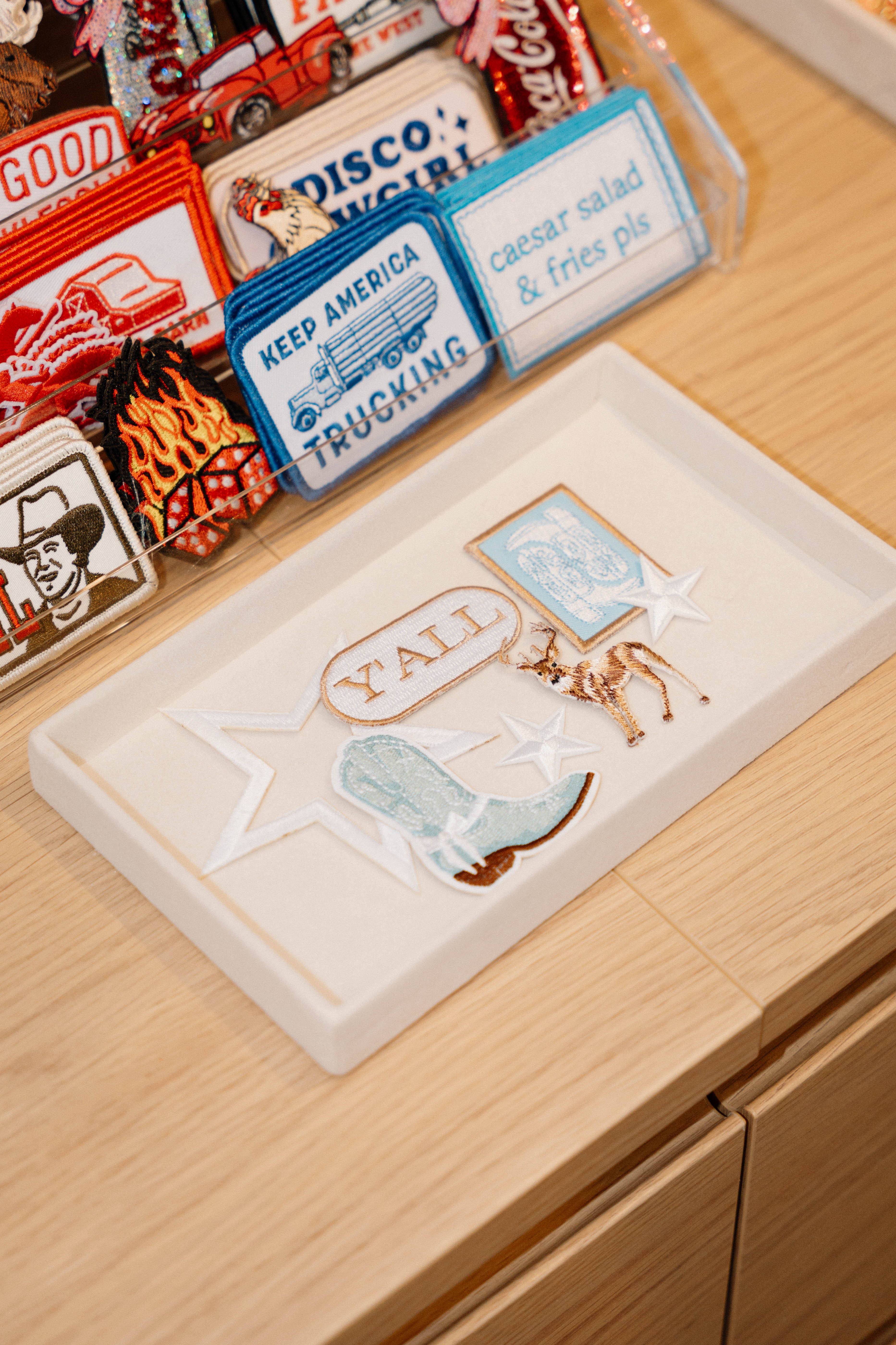 Western-themed embroidered patches on a light wood counter — “Y’ALL” speech bubble, turquoise cowboy boot, pronghorn and star patches in a white tray, next to colorful trucker and novelty patches on display.