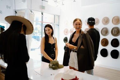 Shoppers laughing and trying on hats in a bright, modern hat boutique — a smiling woman in a blazer and white shorts chats by a counter while a wall of neutral-tone fedoras is on display.