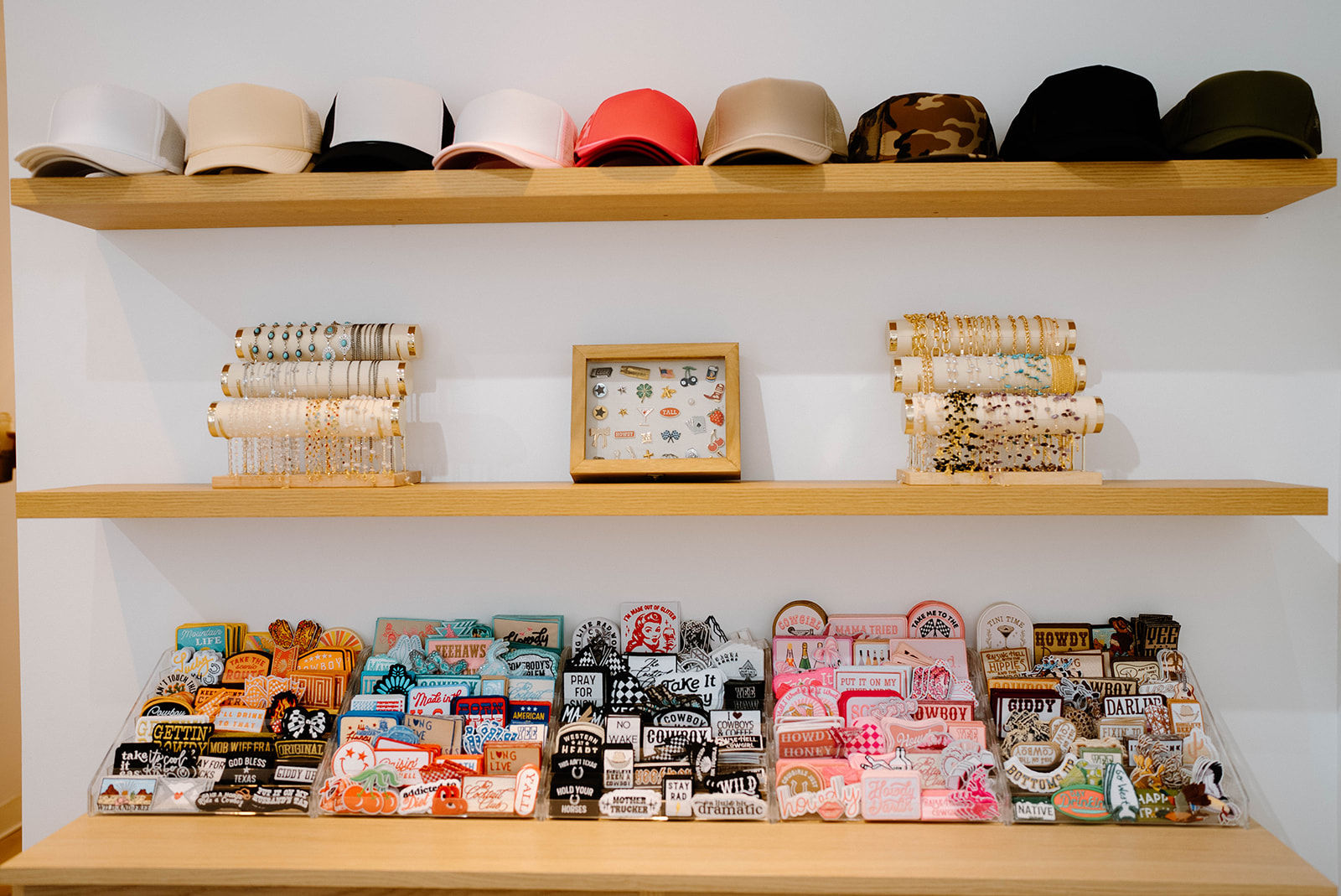 Wooden wall shelves in a boutique displaying neutral and camo caps on the top shelf, rows of gold and beaded bracelets with a framed pin board on the middle shelf, and colorful stickers and embroidered patches neatly organized on the bottom shelf.