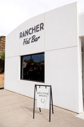White stucco storefront of a hat boutique with bold black signage and a sidewalk A-frame sign that reads “Welcome Brides” with a horseshoe graphic.