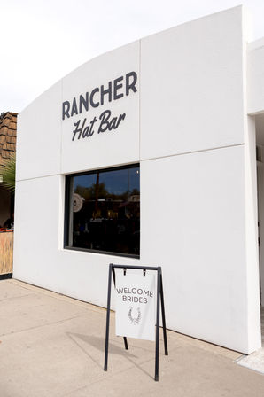 White stucco storefront of a hat boutique with bold black signage and a sidewalk A-frame sign that reads “Welcome Brides” with a horseshoe graphic.