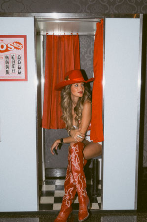 Woman in a red cowboy hat and tall embroidered red boots posing inside a vintage photo booth with red curtains and black-and-white tiled floor
