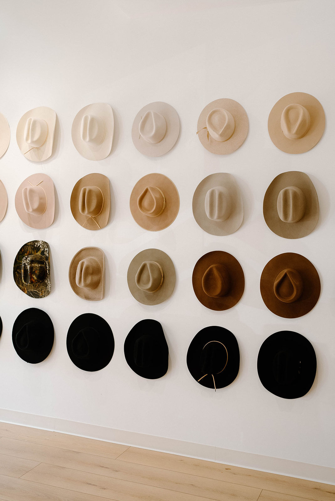 Stylish wall display of neutral wide‑brim felt hats arranged in rows from cream to black in a minimalist boutique interior with light wood flooring.
