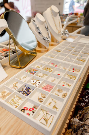 Boutique jewelry display with organized tray of gold earrings, charms and colorful pins, mirror and necklace busts on a wooden retail counter