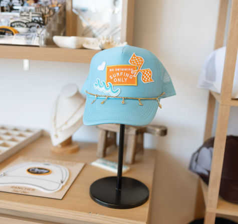Light turquoise trucker hat with orange "No Swimming Surfing Only" patch, wave embroidery and dangling gold chain, showcased on a black stand on a wooden boutique shelf
