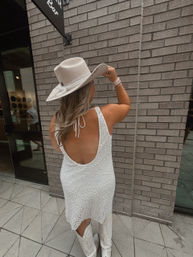 Back view of a person in a white textured mini dress, white cowboy boots and beige wide-brim hat posing against a gray brick storefront on a downtown sidewalk