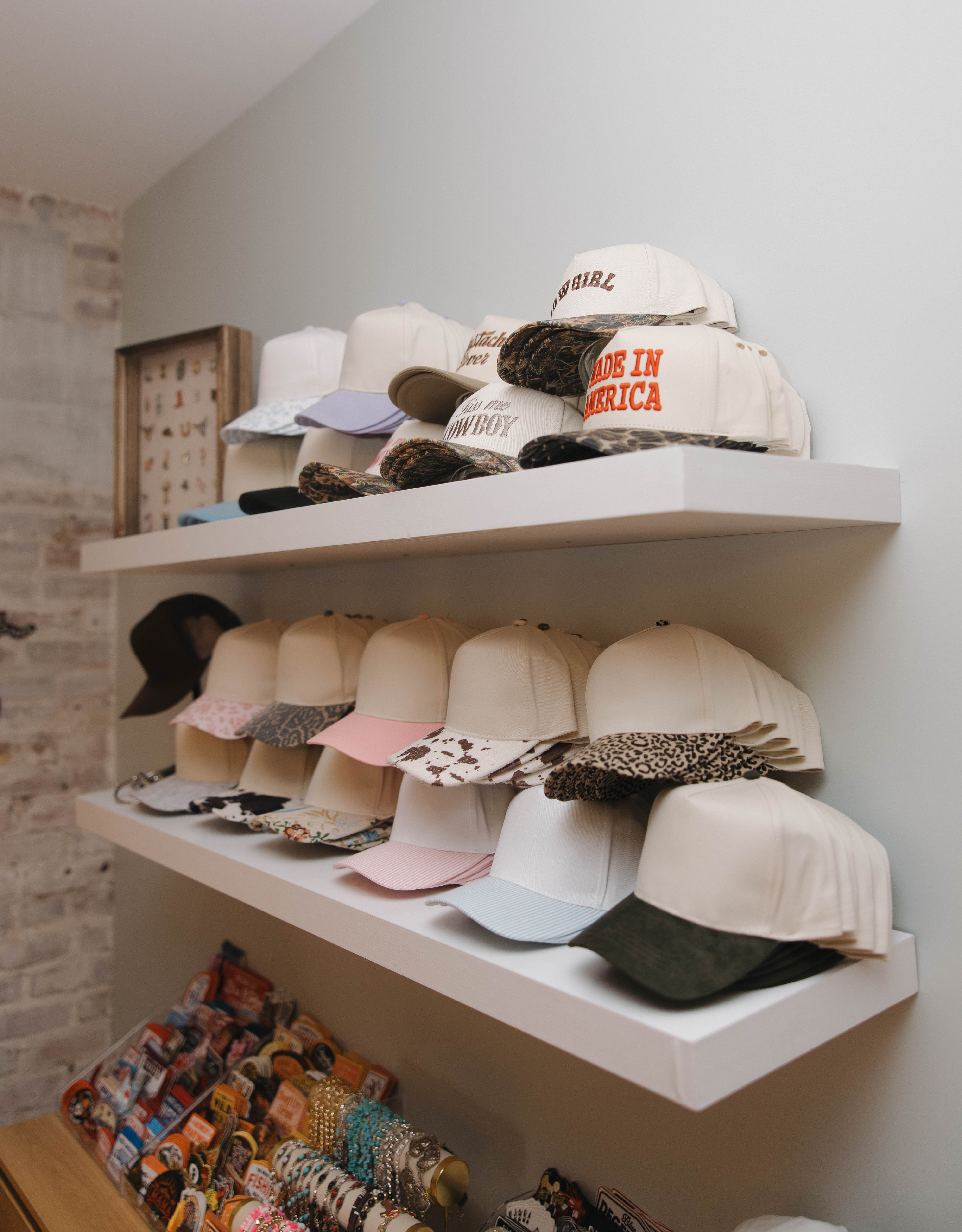 Retail hat display with stacked baseball caps on white floating shelves — pastel, animal-print and slogan caps (including ‘Made in America’), with pins and bracelets below.