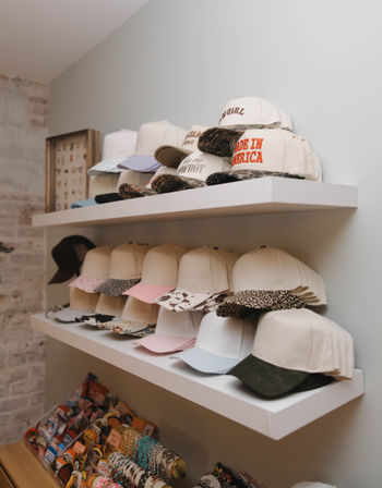 Retail hat display with stacked baseball caps on white floating shelves — pastel, animal-print and slogan caps (including ‘Made in America’), with pins and bracelets below.