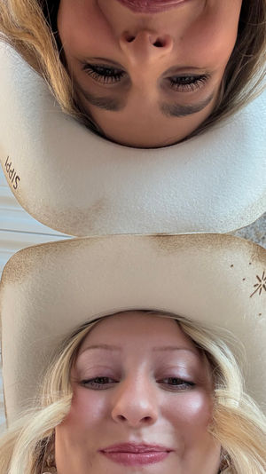 Playful low-angle selfie of two people wearing worn cowboy hats, smiling head-to-head beneath a corrugated metal barn roof