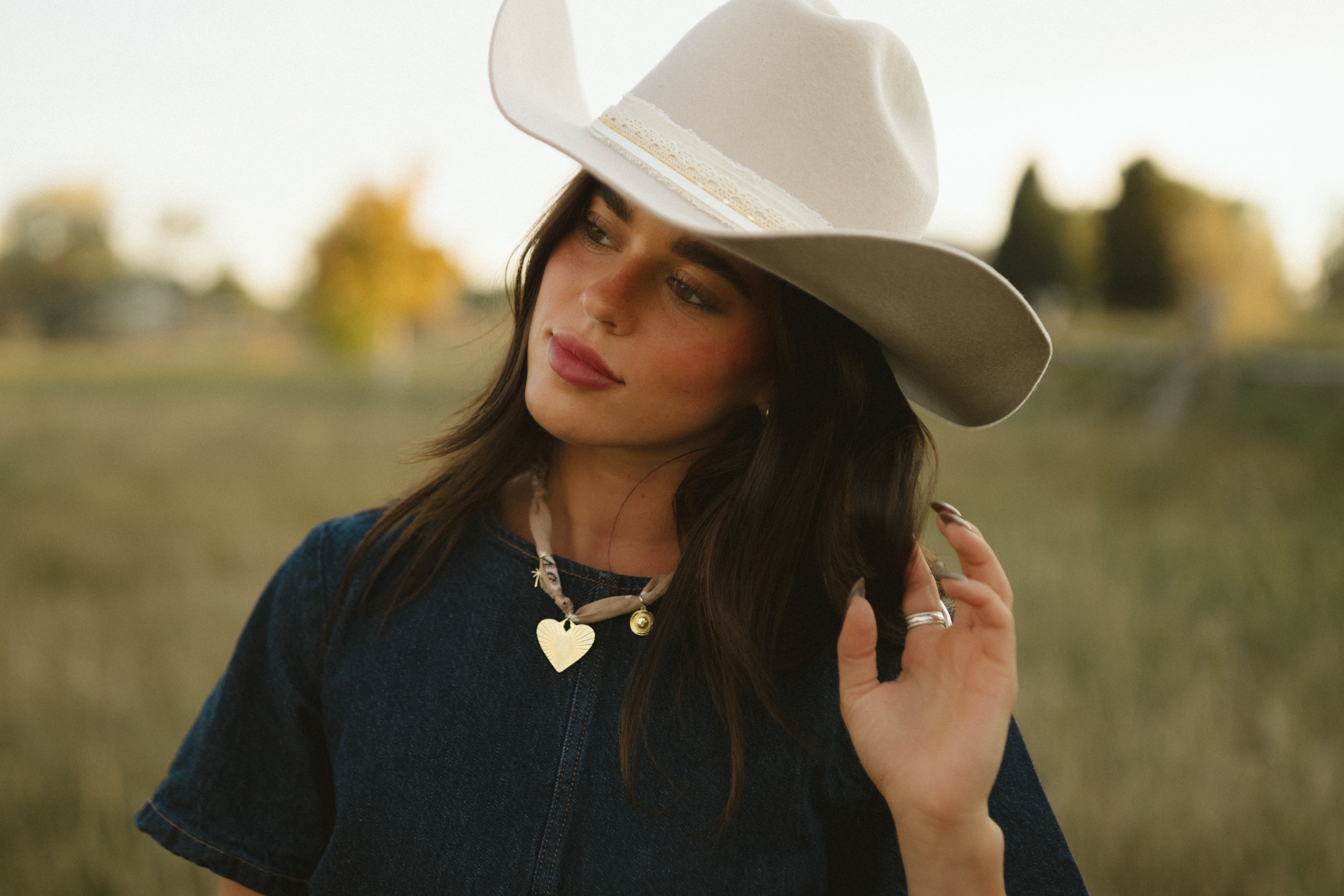 Woman in beige cowboy hat and denim top standing in a rural field at golden hour, touching her hair and wearing a gold heart pendant — relaxed western-style portrait.