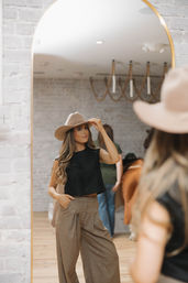 Woman trying on a beige wide-brim hat while checking her reflection in a tall arched mirror inside a boutique with exposed white brick walls, wearing a black cropped top and high-waisted wide-leg trousers.