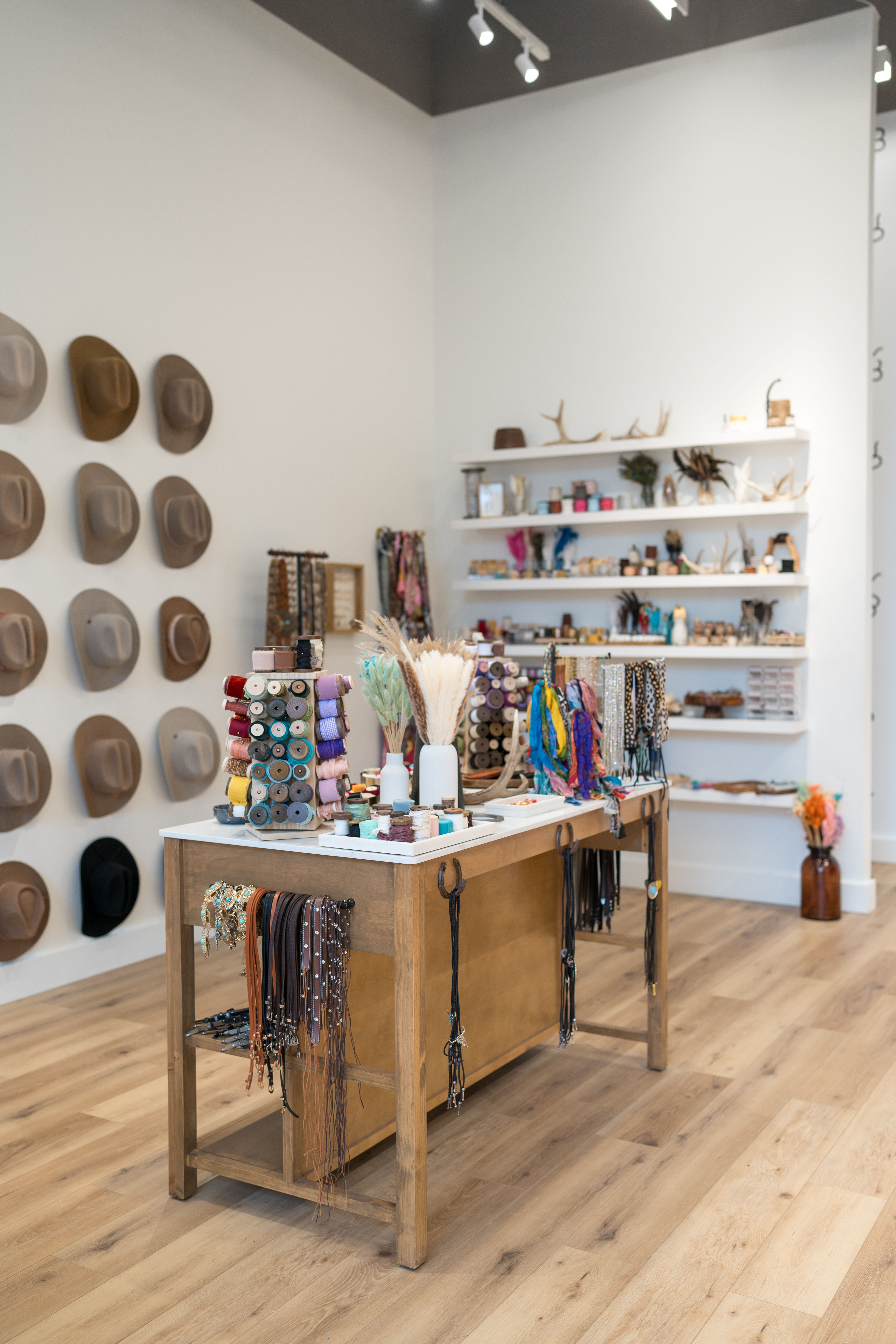 Bright boho accessory boutique interior with a wall of hats, wooden center table displaying colorful ribbon spools, leather belts, necklaces and craft-filled shelves on light wood floors