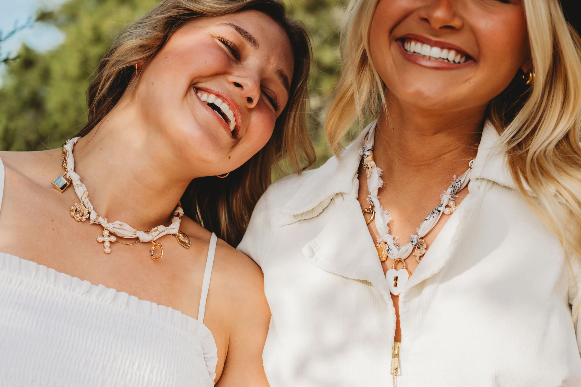 Close-up of two smiling people outdoors wearing white tops and braided charm necklaces, summer fashion accessory.