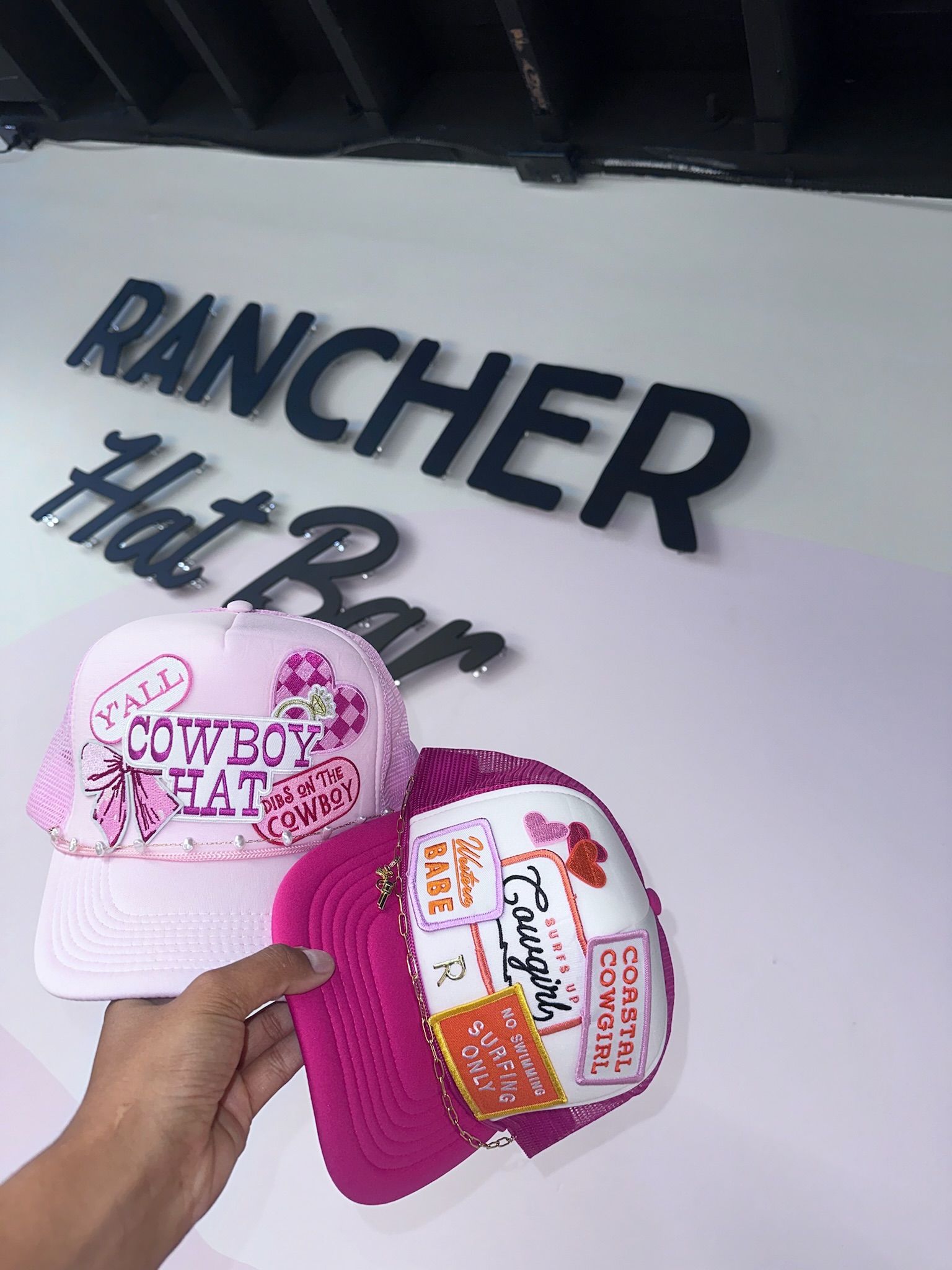 Hand holding two pink embroidered trucker hats with cowboy and cowgirl patches, hearts and chain accents, photographed inside a hat shop under a black wall sign.