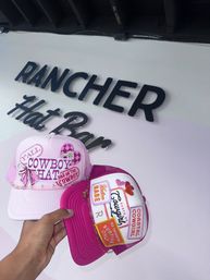 Hand holding two pink embroidered trucker hats with cowboy and cowgirl patches, hearts and chain accents, photographed inside a hat shop under a black wall sign.